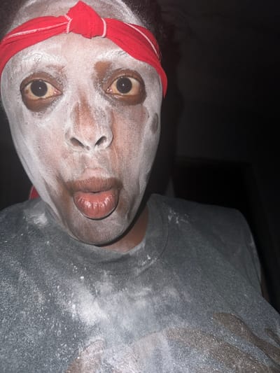 The image shows a close-up, front-facing view of a single individual located in Sabon Tasha, Nigeria. The subject, an adult with dark skin, has their face heavily coated with a white powdery substance, which extends onto their neck and a dark grey t-shirt. A red fabric headband is worn across the forehead. The subject's eyes are wide open, with dilated pupils and light brown sclera. The mouth is slightly open, with lips puckered forward, and the tip of the tongue is partially visible. The t-shirt is also dusted with the white powder. The background is dark and indistinct, indicating low-light conditions or an interior setting.