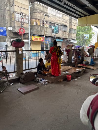 The image depicts a street scene in Kolkata, India. A group of people are gathered on a sidewalk next to a metal railing. One woman is wearing a red and yellow sari. Several men and boys are seated or squatting on the ground. Debris and trash are scattered around them. In the background, a multi-story building shows signs of disrepair, with exposed electrical wiring and faded advertisements. Signage in Bengali is visible, as well as a poster for "prayag." The scene appears to be a lower-income area with signs of poverty and street life.