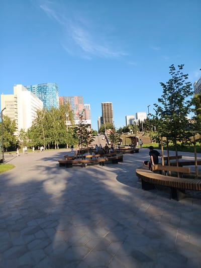 The image captures a modern urban park setting under a clear blue sky. In the background, a cluster of contemporary high-rise buildings dominates the skyline, featuring a mix of architectural styles and colors, including glass facades and visible concrete structures indicating ongoing construction. The foreground is a paved area with an intricate geometric pattern, shaded by scattered trees. Several curved wooden benches are strategically placed throughout the park, some occupied by individuals. One person, wearing a dark t-shirt and shorts, is seated on a bench in the middle right of the frame, facing away from the camera. Other people are visible walking in the mid-ground. Lush green trees and grass add a natural element to the urban landscape. The scene appears to be a public space designed for relaxation and recreation, possibly in the city of Novosibirsk, Russia, given the architectural style of the buildings.
