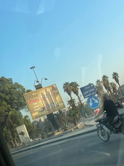 The image depicts a daytime street scene in Ash-Shaghour Municipality, Syria, captured from inside a moving vehicle. The sky is clear blue with a slight gradient towards a lighter hue near the horizon.

In the mid-ground, a dark-clothed individual rides a black motorcycle from right to left on a paved road. To the left of the motorcyclist, a blue rectangular road sign with white Arabic text reads "مركز انطلاق الجنوب باب مصلى" (Southern Launch Center / Bab Musalla), with a right-pointing arrow. Below this, a circular blue sign shows two horizontal white arrows pointing left and right, indicating a two-way street or diverging traffic. A dark-colored car is partially visible in the distant background, to the right of the signs.

On the left side of the road, a large billboard displays an advertisement for "Oliva" brand hair products, featuring three bottles of shampoo or conditioner. Arabic text on the billboard translates to "New from Oliva Keratin Shampoo: Strength, Shine, Softness." Below and slightly behind the billboard, a tall streetlight pole with multiple luminaires extends towards the sky. Further left, a dense cluster of green trees and a white, angular stone monument or sculpture are visible.

Numerous tall palm trees line the road in the mid-ground and background, interspersed with other smaller green foliage and some agave-like plants. Distant residential buildings are partially obscured by the trees and billboards, indicating an urban environment. The foreground shows reflections on what appears to be a vehicle's windshield.