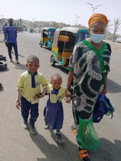 This image captures a street scene in Mumbwa, Zambia, under bright, hazy daylight. In the foreground, three individuals walk from right to left on an asphalt road. On the right, an adult female wears an orange headwrap, a light blue surgical mask, a dark green patterned dress with green fringe at the hem, and orange and black sandals. She carries a blue backpack with green accents in her left hand and appears to be holding the right hand of a smaller child. Her abdomen is distended.
To her left, a young child, possibly a girl, wears a yellow collared shirt with a blue bow tie and a school emblem, a dark blue overall-style garment, striped socks, and dark lace-up shoes. This child holds the hand of the adult female and the left hand of an older boy to its left. The older boy wears a yellow collared shirt with a blue tie and a school emblem, a striped long-sleeved undershirt, dark blue trousers, and grey lace-up shoes.
In the midground, to the left of the walking group, an adult male stands on the roadside, dressed in a purple short-sleeved shirt and dark trousers. Several discarded black tires lie on the ground near his feet. Behind the main subjects, a line of three-wheeled auto-rickshaws (tuk-tuks), primarily light blue/green and yellow, are visible on the road. One vehicle has "PFA 9004" visible on its side. The background features a hazy sky, distant hills, sparse vegetation including palm trees, and modern streetlights along the road. Various distant structures are also visible.