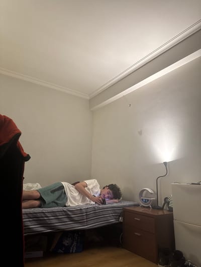 A young man with curly dark hair is standing in a bedroom, looking at his phone. He is wearing a white t-shirt and green shorts. He is leaning against a striped bed frame. To his right is a dark wood nightstand with two drawers and a lamp. On top of the nightstand are shoes. The room has white walls and ceiling with decorative molding. A desk lamp is clipped to the top of the nightstand. The location is likely inside a house or apartment, possibly in Bragança, Portugal, given the presence of the game "Carragadinha de Vantagens" which is a game sold by the Lidl chain of stores in Portugal.