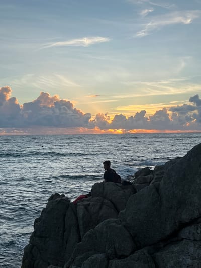 A person sits on a rocky shore overlooking a body of water at sunset. The sky is filled with colorful clouds, illuminated by the setting sun. The water is choppy, with small waves breaking against the rocks. The person is silhouetted against the sky, making it difficult to discern their features. A red object, possibly a backpack or clothing, is near the person. The scene appears to be a natural landscape, possibly a beach or a coastal area, given the presence of rocks and the ocean. The image has a serene and tranquil atmosphere, typical of a sunset scene.