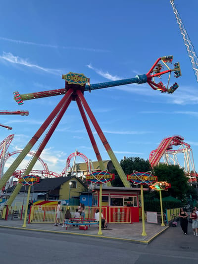 The image captures an amusement park scene with a vibrant, towering ride as the central focus. This ride features a tall, blue, LED-lit pole from which four large, red arms extend outwards, each supporting a yellow gondola filled with passengers. The gondolas appear to be spinning or swinging, suggesting the ride is in motion. In the background, a red roller coaster with white supports winds through the park, and another, smaller ride with a blue and yellow structure is visible to the left.
The setting is an outdoor amusement park under a clear blue sky with wispy white clouds. The time of day appears to be late afternoon or early evening, as indicated by the warm, soft lighting. People can be seen in and around the rides and in a waiting area near a building with a red and yellow facade. Some individuals are seated in the ride's gondolas, while others stand in queues or stroll through the park.
The prominent ride has a sign that reads "DISCOVERY" in large, colorful letters, embellished with atom-like graphics. Another sign on the same ride displays the word "VOLTIGE". The overall atmosphere seems energetic and fun, with the bright colors of the rides and the clear sky contributing to a cheerful mood. The location cues suggest this is an amusement park in Vienna, Austria.
