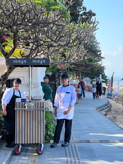 This media file depicts a scene on a beach in Denpasar, Indonesia. The foreground features a wheeled cart with a striped wooden exterior, positioned next to a tree with bare, intricate branches and some green leaves. Two individuals, dressed in white chef coats and black caps, stand near the cart. One is closer to the camera, facing slightly away, while the other is further back.

In the mid-ground, a group of people are visible, some standing and others seated. They are near a beach umbrella and a food stall. The background reveals a sandy beach with more umbrellas and lounge chairs, leading to the ocean with calm water. The sky is clear and blue, suggesting a sunny day. The overall atmosphere appears relaxed and casual, typical of a beachside location.

Visible text on a black sign attached to the tree reads "maya maya" in a stylized font, repeated vertically. Another sign on a white structure behind the cart also has text, though it is mostly obscured and illegible. The time of day appears to be late morning or early afternoon, given the bright sunlight and shadows.