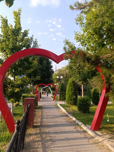 This media file captures a park scene in Fergana City, Uzbekistan, likely during the late afternoon or early evening due to the long shadows cast across the paved pathway. The weather appears clear with a few wispy clouds dotting the bright blue sky.
The dominant subjects are two large, vibrant red heart-shaped archways situated along a walkway. The foreground archway is prominent, framing the path as it recedes into the distance. Further along the path, a second, smaller red heart archway is visible, creating a sense of depth and perspective. Both arches appear to be decorative installations within the park.
The setting is a well-maintained park with lush green trees and manicured bushes lining the path. A black wrought-iron fence with decorative finials runs along the left side of the walkway, alongside a low brick wall. Several people are present, walking along the path. In the middle distance, two individuals are seen walking, one seemingly riding a scooter or bicycle. Their presence suggests the park is a recreational space.
Notable details include the sunlight illuminating the scene, creating a warm glow and distinct shadows. The overall atmosphere is peaceful and inviting, characteristic of a public park designed for relaxation and enjoyment. No visible text is present in the image.