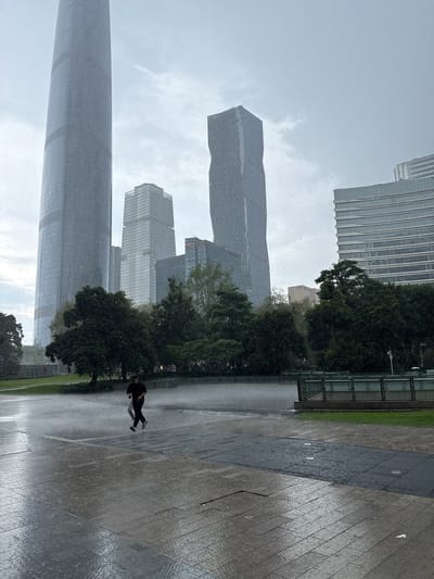 The image depicts a cityscape in Tianhe District, China, during a rain shower. Several modern skyscrapers are visible in the background, including a tall, cylindrical tower on the left and a twisted, angular tower in the center. In the foreground, two individuals are running on a wet, paved plaza. Trees and green spaces are situated between the plaza and the buildings. The rain is visible as streaks and droplets in the air, and the ground is reflecting the ambient light due to wetness. The sky is overcast, contributing to a muted color palette.