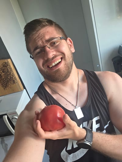 In the city of Yagodovo, Bulgaria, a male individual is captured in a brightly lit indoor environment, likely a kitchen or dining area. The man, appearing to be in his 20s or 30s, has short light-brown hair, a well-trimmed beard, and is wearing dark-framed eyeglasses. He is smiling broadly, revealing his teeth, and is looking directly at the camera, suggesting a selfie. He wears a dark tank top, part of a white "NIKE" logo visible on the chest. A silver-colored chain necklace with a blank, rectangular silver pendant hangs around his neck. On his left wrist, he wears a smartwatch with a black strap, displaying "HUAWEI WATCH" text. His left hand is holding a single, round, ripe red tomato. In the background to the left, a light-colored wooden cutting board with an engraved design featuring grapes and a barrel is visible, resting on a white "OLIMPIC" brand microwave oven. The surrounding environment consists of light-colored walls and surfaces, implying a modern and clean setting.