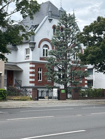 The image shows a street view of a multi-story building with a red brick and white stucco facade, located in Berlin, Germany. The building has arched windows on the upper floors, decorative turrets on the roof, and a prominent tree in front. A paved sidewalk and a street with white lane markings are visible in the foreground. The sky is overcast.