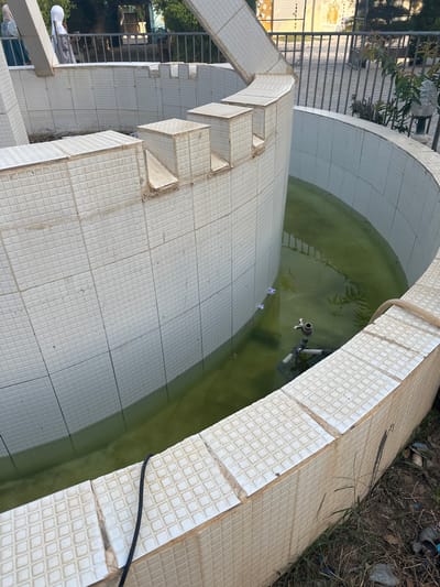 The image shows a circular, tiled fountain in an outdoor urban setting, possibly within Batagarawa, Nigeria. The fountain's basin is partially filled with murky green water, and a submersible pump with piping is visible in the water. The white tiles on the fountain's exterior and interior appear aged, with some signs of wear and tear. A light-colored hose snakes out of the fountain and onto the ground. In the background, a metal railing separates the fountain area from a paved walkway. Beyond the railing, there are trees and some buildings or structures, including a mural with figures on a wall. Two individuals, one wearing a hijab and the other a light-colored headscarf, are visible in the background near the railing. The ground surrounding the fountain is rough, with dirt, dry grass, and some litter.