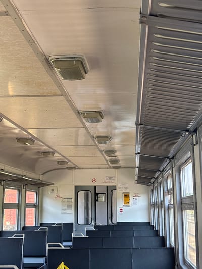 The image depicts the interior of a train car. The perspective is from a low angle, looking forward along the length of the carriage. The ceiling is a light, textured material with several rectangular light fixtures recessed into it. Along the right side, there are rows of windows, each divided into multiple panes, with a metal grate-like structure running above them. The seats are upholstered in dark fabric and appear to be arranged in a bench-style configuration.
The walls are white, and there are various signs posted. One sign with a red number "8" is visible on the back wall near a doorway. Another sign, predominantly yellow with text and icons, is mounted on the wall. There are also smaller notices with Cyrillic text. The floor is not clearly visible. The environment appears to be utilitarian and functional, typical of public transportation. No people are visible in the image. The setting is likely within a train car on a route that may pass through or originate from the Ганчарскі сельскі Савет region in Belarus, though the image itself does not provide direct evidence of this specific location.