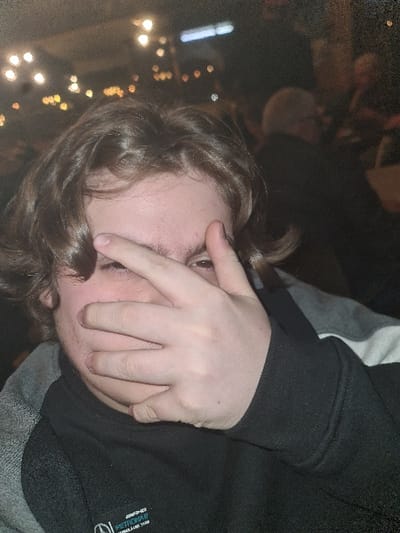 A young person covers their face with their hand, peering out from between their fingers. They have light skin, light brown hair, and are wearing a black and grey hooded sweatshirt. The background is dimly lit, with bokeh lights visible, suggesting an indoor setting. The overall image has a slightly blurred quality, possibly due to low light conditions.