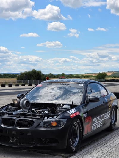 A black, modified BMW E92 M3 drag car is parked on an asphalt track. The hood is open, revealing a large turbocharger and engine components. The car has racing decals, including "DINOSAUR TEAM" and "PERFORMANCE SERVICE". The headlights have an orange, reptilian eye design. The background shows a grassy field, trees, and a cloudy blue sky. A digital timer displaying "8.8.2.658" is visible in the distance. This scene appears to be at a drag racing event. The location is not specified as Akatsievo, Bulgaria.