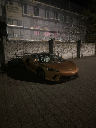 The image displays a metallic bronze convertible sports car, identified as a McLaren, parked on dark grey rectangular block paving at night in Dietlikon, Switzerland. The vehicle is angled towards the right, featuring a black soft top or hardtop and visible light-colored (white/light grey) interior seating. Faint orange light emanates from the side mirror areas.

Behind the vehicle, a light-colored brick wall adorned with bare, leafless vines extends horizontally, punctuated by vertical stone pillars. Intense artificial illumination from the left casts sharp shadows of the car and the vines onto the wall.

In the background, a multi-story building is under construction or renovation, entirely covered by scaffolding and white protective netting. A large white banner attached to the scaffolding's upper left section reads "SICHERHEIT. FLEXIBILITÄT. GERÜSTBAU." and "HUBER GERÜSTBAU SCHAFT," with the Swiss phone number "044 941 50 80." Rectangular areas of pink light are visible through the building's netting, suggesting internal illumination. Leafless trees are visible beyond the wall on the right. No individuals are present in the scene.