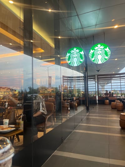 The image captures the interior and exterior view of a Starbucks coffee shop in Dumai City, Indonesia, seen through large glass windows. Inside, several customers are visible. In the foreground, a person is seated in a brown leather chair, wearing a patterned top and dark pants, holding a shopping bag. To the left, a table holds a white cup with a lid and straw, an iced beverage with a straw, and a plate with a croissant.
Further inside the establishment, a man in a brown polo shirt is seated on a wooden stool, interacting with a small item on a table. In the background, wooden barrel-shaped seats are arranged, and large windows offer a view of the city's skyline, characterized by various buildings, including what appears to be a lighthouse or tower on the right. Two prominent green Starbucks circular logos with the siren image are suspended from the ceiling. The overall ambiance suggests a modern, well-lit coffee shop with a blend of indoor seating and an outdoor view, reflecting the urban environment of Dumai City. The lighting appears to be a mix of artificial interior lights and natural light from the dusk sky, casting reflections on the glass surfaces.
