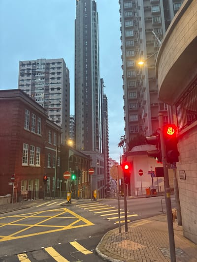 The image depicts a street scene in Hong Kong, China. The view is from a low angle, looking up at tall buildings. The buildings are typical Hong Kong residential towers, with many windows and air conditioning units. In the foreground, there is a red brick building. The street has a yellow box junction marked on it. There are traffic lights showing both red and green signals. Street signs are also visible. The sky is overcast. There are no people visible in the frame.