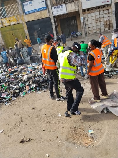 The image displays a daylight scene on a dusty, unpaved street in Fagge C, Kano State, Nigeria, specifically along Kano Street as indicated by signage. A large, sprawling accumulation of mixed refuse, including plastic bags, fabric, and various debris, occupies the left side of the midground. Multiple individuals are present, many actively engaged in or observing a cleanup operation.
In the foreground and midground, several individuals are prominent. A man wearing a neon green high-visibility vest, a black cap with "RATEL" visible, black trousers, and striped sandals is bending forward. To his immediate left, another man in an orange high-visibility vest, black shirt, and black track pants with green stripes stands with his back partially to the camera. To the right of the man in the green vest, a woman wearing an orange high-visibility vest, a black shirt, brown trousers, and a black face mask stands, facing the man in the green vest.
In the background, additional individuals are clustered around the refuse pile, some also wearing orange high-visibility vests and seemingly handling waste, with one individual on the far right holding a tool resembling a shovel or broom. Other individuals are visible in the distance, some standing, one seated on a low structure. The background consists of multi-story buildings with partially obscured or closed ground-floor storefronts. Visible signage includes "NEWCLIME / 90-GOMBO ELECTRICAL STORE" and "ONYICK INDUSTRIAL COMPANY / ... Kano Street, Fagge, Kano State." The scene depicts a community cleanup effort.