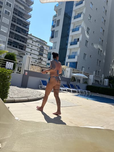 A woman, viewed from behind, stands on a tiled patio beside a swimming pool. She is wearing a leopard-print bikini and has visible tattoos on her back, legs, and arms. She is holding a black object in her right hand. In the background, there are several modern apartment buildings, some with blue-tinted windows and balconies. There are lounge chairs and an umbrella near the pool, and a green hedge to the left of the frame. The scene is illuminated by bright sunlight, casting shadows on the ground. The location is Alanya, Turkey.