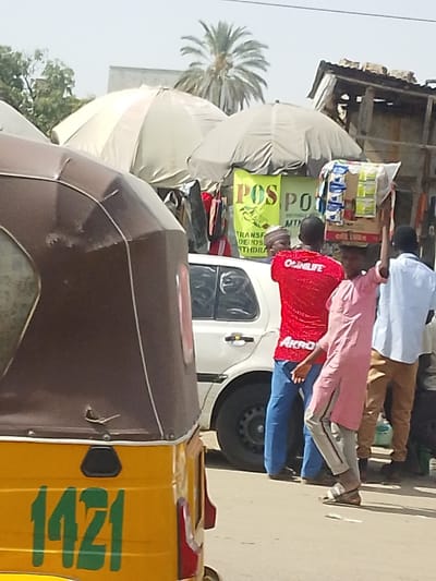This image captures a street scene in Fagge A, Nigeria. In the foreground, the rear section of a yellow and brown three-wheeled auto-rickshaw is visible, bearing the green numbers "1421". To its right, a white four-door sedan is parked or stationary. Several individuals are present: a person in a red shirt displaying "OGUNLIFE" and "AKRO" on the back, wearing blue pants and sandals, stands near the white car, facing slightly right. To their left, another person, dressed in a pink tunic over grey striped pants and sandals, carries a cardboard box laden with small packaged goods on their head, facing left. Further right, a man in a light blue shirt and tan pants is seen from behind. Behind the people and vehicles, two large open market umbrellas, one light beige and one grey, provide shade over market stalls. These stalls display hanging clothing and two bright yellow banners with "POS" prominently printed, indicating "TRANSFER, DEPOSITS, WITHDRAWAL" services. A dilapidated structure with a corrugated roof is visible behind the stalls. In the background, a palm tree and other sparse vegetation are present under a bright, hazy sky.