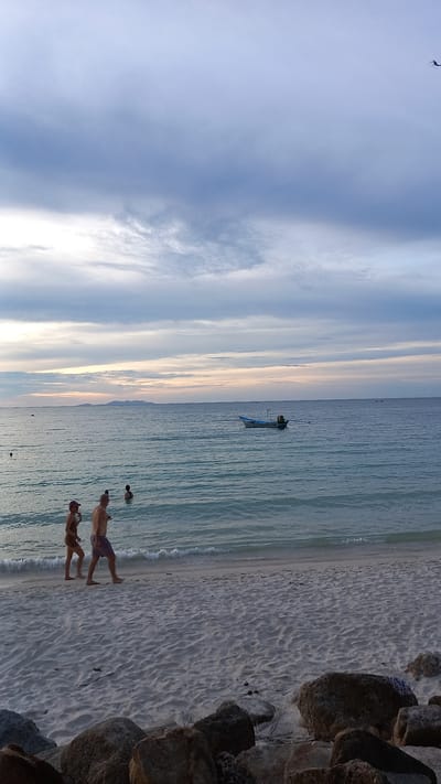This is a photograph taken on the beach in ตำบลเกาะพะงัน, Thailand. In the foreground, there is white sand with several large rocks scattered along the edge of the water. Two people are walking along the shoreline from left to right. The person on the left is wearing a dark bikini and a pink hat. The person on the right is a man wearing dark shorts. Further out in the water, there are a few more people swimming. A small blue and white boat with an outboard motor is anchored in the middle distance. In the far distance, a faint landmass is visible on the horizon. The sky is filled with dramatic clouds in shades of gray and white, with hints of pastel colors near the horizon, suggesting either sunrise or sunset. A small dark object, possibly a bird or a drone, is visible in the upper right corner of the sky. The water is calm with gentle waves lapping at the shore.