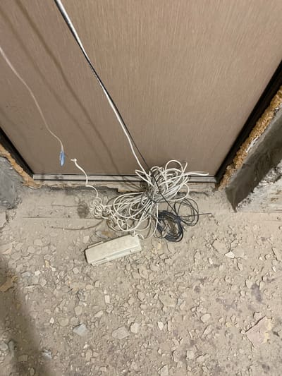 This media file depicts a close-up, eye-level view of construction debris and electrical wiring next to a door frame. The scene is set outdoors or in an unfinished indoor space, likely during daylight hours, as indicated by the lighting. The ground is covered in rubble and concrete fragments, suggesting an active construction or demolition site.
The primary objects in the frame are several tangled bundles of electrical wires, one bundle predominantly white and another black. These wires are loosely coiled and appear to be either recently installed or discarded. A white plastic electrical box is visible lying on the rubble. Adjacent to the wires and rubble is the edge of a light brown door, which has a visible seam running down its center. The door frame itself is lined with expanding foam insulation, a common material used in construction to seal gaps.
The overall impression is one of ongoing work, with materials and tools laid out or abandoned. There are no people visible in the image. The location context of Ulyanovsk, Russia, suggests this is a scene from that region. The image is factual and devoid of any discernible text. The focus is on the raw, unfinished state of the area and the presence of electrical components.