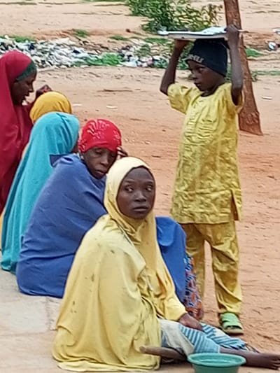 The image shows a group of people, likely women and children, in an outdoor setting that appears to be a dusty, open area. The people are wearing traditional clothing, with headscarves and brightly colored fabrics in shades of red, yellow, blue, and green. In the foreground, a person in a yellow headscarf is seated, with their legs extended and a wooden stick nearby. A green bowl is also visible in the foreground, partially obscured by the person. To the left, a person in a red headscarf is also seated. Further back, several other individuals are positioned, including a child wearing a yellow outfit and a dark beanie, carrying a tray on their head. In the background, there are piles of debris, possibly trash, and sparse vegetation. The ground is sandy and uneven. The overall impression is a snapshot of daily life in a community. The image, with its focus on traditional attire and the presence of children, along with the rustic environment, is consistent with scenes found in various communities within Gombe State, Nigeria.