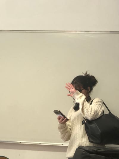 The scene is an interior location in Schiltigheim, France, characterized by a large white board against a light-colored wall, consistent with a classroom or meeting room. A female individual is captured from the mid-torso upward, positioned facing the viewer. Her dark, curly hair is gathered into a messy bun, and she wears eyeglasses. She is dressed in a cream-colored cable-knit sweater, with a dark accessory, possibly headphones, visible around her neck. A metallic ring adorns the fourth finger of her left hand. She holds a dark-cased smartphone with a white attachment on its reverse side in her left hand, directed downwards. Her right hand is raised, palm open, positioned in front of her face, partially obscuring her facial features. A large black shoulder bag is carried on her right side. Partial views of a brown, possibly wooden, chair are visible in the lower left, and a dark bag or object is present in the lower right portion of the frame.