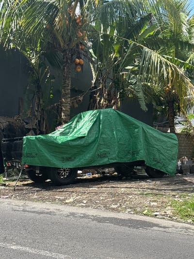 In Tegallalang, Indonesia, a utility vehicle is parked under a bright green tarp, its entirety obscured except for a section of the rear bed. The vehicle, likely a pickup truck or similar model, has a license plate that reads "DK 8154 KU". The tarp displays a repeating logo with the text "SUPER KOMODO". The vehicle is situated on a gravelly, uneven surface adjacent to a paved road. Behind the vehicle, tall palm trees with visible coconuts and large leafy plants create a dense, tropical environment. The background reveals a dark building wall and some construction materials, including a stack of bricks and pipes, suggesting a semi-rural or developing area. The lighting indicates it is daytime, with shadows cast on the road, implying the sun is at a moderate angle. There are no people or animals visible in the scene.