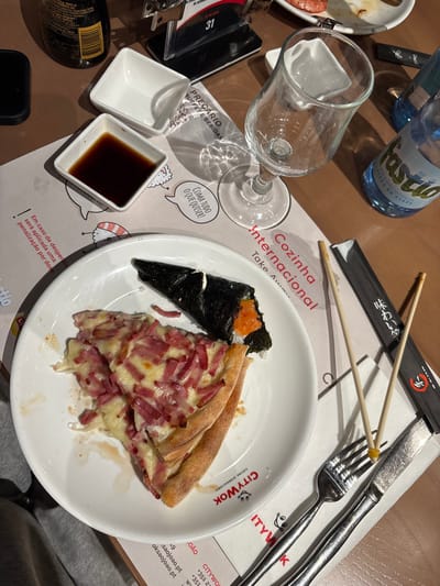 The image captures a table setting at a restaurant, specifically the "City Wok Cozinha Internacional" establishment in Vila Nova de Gaia, Portugal. A plate holds a slice of pizza with ham and cheese, alongside a temaki (seaweed cone) filled with rice and salmon roe. Nearby, a small dish contains soy sauce. Chopsticks rest beside a fork and knife, indicating that both Asian and Western cuisines are available. A nearly empty water bottle and an empty wine glass are also present. The tablecloth is printed with menu information in Portuguese, mentioning that a penalty will be applied for food waste. The restaurant's branding is prominently displayed on the plate.