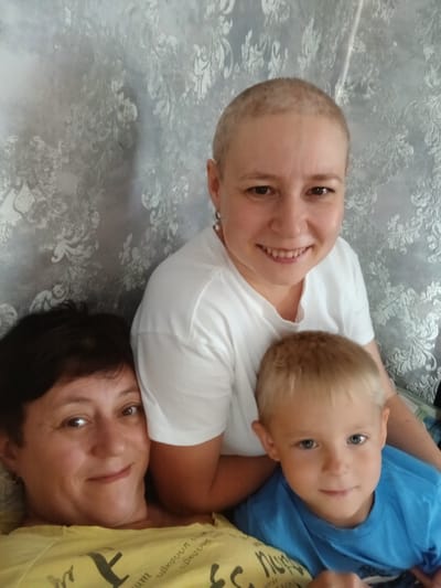 Three people are posing for a selfie indoors. The foreground is occupied by two individuals, a woman with short dark hair and a boy with blond hair. The woman is lying down, her head resting on a yellow shirt with some text visible. The boy is wearing a blue shirt and is positioned in front of the woman. Behind them, a woman with a shaved head is smiling and looking at the camera. She is wearing a white t-shirt and is positioned slightly higher than the other two. The background consists of a textured wallpaper with a floral pattern. The overall lighting suggests an indoor setting, possibly a living room. The location is identified as Kolyvan, Russia.