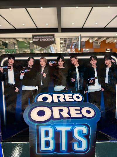 The image shows a promotional display for Oreo BTS cookies, featuring life-sized cardboard cutouts of seven men, presumably members of the K-pop group BTS. They are dressed in dark clothing and holding Oreo products. The display is positioned in front of a blurred background that appears to be an indoor retail environment with ceiling lights visible. A sign with Thai text and "SELF CHECKOUT" in English is visible in the upper portion of the image, suggesting the location is a store or supermarket. The overall impression is a vibrant commercial setting.