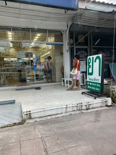 A person wearing an orange shirt and patterned shorts stands on a sidewalk in front of a pharmacy. They are holding a white tote bag over their shoulder and appear to be looking down at their hands. The pharmacy has a large glass storefront with displays of products visible inside. A green and white sign above the entrance reads "ยา" (Ya) which means medicine in Thai, and also "โดยเภสัชกร" (Doi Phenachakorn) meaning "By Pharmacist". Below this, it lists operating hours as "09:00-21:00 น." and a phone number. Another sign on the storefront says "WELCOME OPEN 09.00-21.00 น.". The scene is set in an outdoor environment with a concrete sidewalk and a building facade. The lighting suggests it might be daytime. The overall atmosphere appears to be casual and everyday. The location is identified as Baan Bo Phut, Thailand, with text on a sign confirming "ต.บ่อผุด อ.เกาะสมุย จ. สุราษฎร์รานี 84320" which translates to Bo Phut Subdistrict, Koh Samui District, Surat Thani Province.
