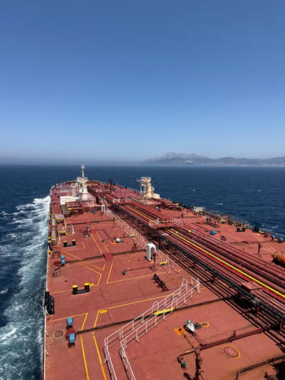 The media file displays a large, red oil tanker sailing on a vast expanse of deep blue ocean. The perspective is from a high angle, looking down and slightly forward onto the deck of the ship. The deck is covered with a network of pipes, many of which are colored red, yellow, and black, indicating different functions. White ladders and railings are visible, along with various equipment such as tanks, hatches, and what appear to be cargo containers.

In the distance, a hazy, mountainous coastline is visible, suggesting the ship is near land. The sky above is a clear, bright blue, transitioning to a lighter shade near the horizon, indicating it is likely daytime, possibly late morning or early afternoon. The sea is moderately rough, with white waves cresting and trailing behind the ship, showing it is in motion. The overall scene evokes a sense of industrial activity and maritime travel.

There is some visible text on the ship. One sign on the side of the superstructure reads "NO SMOKING." Another circular marking on the deck has text that is too small to be legible in the provided image. There are no people clearly visible in the frame.