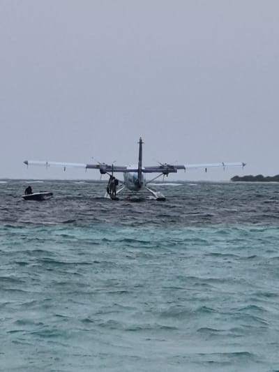 A seaplane, identifiable by its two wing sections, a central propeller, and floatation gear, is floating on the ocean’s surface near Bodufolhudhoo, Maldives. The water shows visible texture indicating possible wave action. A thin strip of land with vegetation appears in the background. The sky is a uniform light gray, suggesting overcast conditions. A person is visible on a board or platform next to the plane.