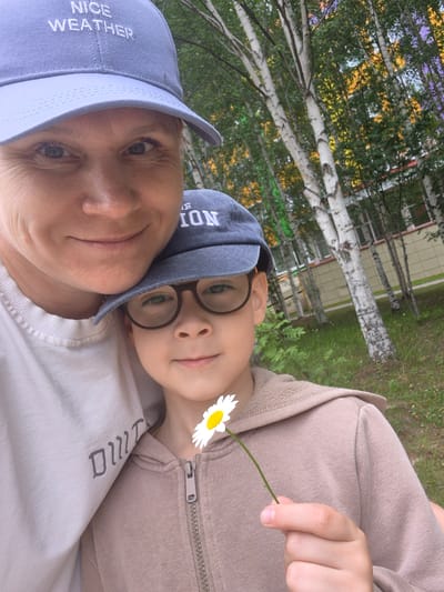 In Nyagan, Russia, a woman and a child are captured in a close-up selfie outdoors. The woman, wearing a light blue baseball cap with "NICE WEATHER" embroidered on it, is positioned in the foreground, her face taking up the left side of the frame. She appears to be smiling, with her eyes looking towards the camera.
Beside her, and slightly behind, is a child wearing a dark gray baseball cap with "ION" visible on it and eyeglasses. The child is holding a single white daisy with a yellow center, presenting it towards the camera with their right hand. The child is also smiling.
The background consists of a natural setting with birch trees, their white bark contrasting with the green foliage. Hints of buildings and possibly painted walls are visible behind the trees. The overall impression is a warm, personal moment shared between the two individuals in a semi-urban, outdoor environment.