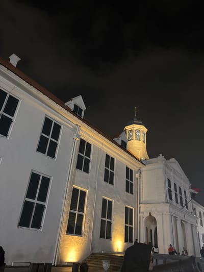 The image shows a night view of the Fatahillah Square in Kota Tua, West Jakarta, Indonesia. The main subject is the historic Dutch colonial building, the Jakarta History Museum (Museum Sejarah Jakarta), formerly known as the Old Batavia Town Hall. The building is illuminated by yellow floodlights, highlighting its white facade and numerous dark-shuttered windows. A prominent tower with a clock face and weather vane stands out against the dark, cloudy sky. In the foreground, there are some dark, indistinct objects that appear to be cannons. Several people are visible on the steps and in front of the building, some sitting and others standing, though their features are not clearly discernible due to the low light and distance. The overall impression is one of a historical landmark at night.