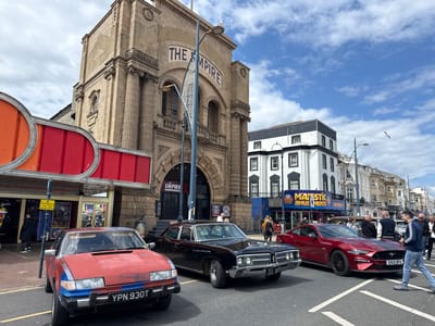 The image showcases a street scene in Great Yarmouth, United Kingdom, featuring three parked cars. In the foreground, a red and blue, somewhat dilapidated Rover SD1 (license plate YPN 930T) is parked on the left, followed by a black 1968 Buick LeSabre (license plate NSC 106F). To the right of the Buick is a red Ford Mustang Mach 1 (license plate EN21 DFU).
Behind the cars is a view of the street and buildings. "The Empire," a large, ornate building, is centered in the background. The building facade features detailed architectural elements and is labeled "THE EMPIRE." To the right of "The Empire" is a multi-story white building with black trim, sporting the sign "MAJESTIC AMUSEMENTS" in blue and yellow.
Several people are visible on the sidewalk and street. A man in jeans and a blue track jacket is walking across the street to the right of the Mustang. Other individuals are seen walking and standing near the buildings in the background. The sky is partly cloudy with some blue patches.