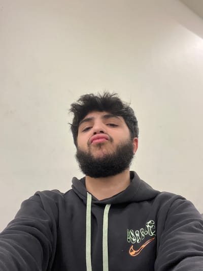 A young man with dark hair and a full beard looks down at the camera. He wears a black Nike hoodie, decorated with pale green and orange accents on the logo and drawstrings. The background is a plain, off-white wall. The image is slightly rotated, giving the impression of looking up at the subject.