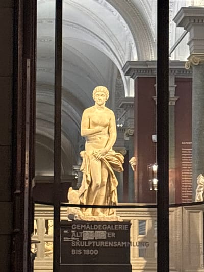 An interior view within the Sempergalerie, part of the Zwinger Palace complex in Dresden, Germany, displays a prominent, light-colored classical marble or stone statue of a female figure. The statue, depicting a nude subject with curly hair, holds her left arm across her chest and her right hand manipulates drapery to cover her lower body. She stands on a stone pedestal. The background reveals a grand hall with high, vaulted ceilings featuring intricate white architectural moldings, arches, and cornices. Columns and pilasters with capital details are visible, alongside walls painted in dark reddish-brown and lighter tones. Artificial illumination brightens the scene. In the lower foreground, a dark rectangular sign with white text identifies "GEMÄLDEGALERIE," "ALTE MEISTER" (partially visible), "SKULPTURENSAMMLUNG," and "BIS 1800," indicating the collection's scope. A red information panel with the German word "KUNST" (art) is partially visible on the right wall. The image is framed by dark vertical and horizontal structural elements, suggesting the vantage point is through an opening or display barrier. No individuals are present.