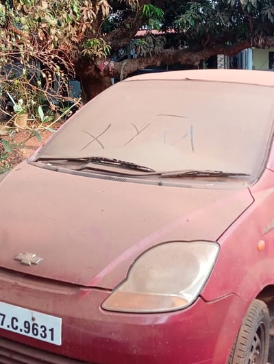 The image displays a dusty red Chevrolet car parked outdoors in what appears to be a residential or semi-rural setting in Arambol, India. The car is heavily coated in a layer of reddish-brown dust, particularly noticeable on its hood, windshield, and headlights, indicating dry and possibly windy conditions.

On the dust-covered windshield, the letters "XYU" have been visibly finger-traced. The front license plate is partially visible at the bottom left, displaying the alphanumeric sequence "7.C.9631". The Chevrolet logo is discernible on the car's hood.

In the background, a large tree with abundant green and brownish foliage dominates the upper portion of the frame, suggesting a tropical environment. Patches of smaller green plants and some branches are visible around the car. A glimpse of a building with light-colored walls can be seen through the tree branches. The bright lighting suggests it is daytime. No people or active events are depicted; the scene focuses on the stationary, dust-covered vehicle.