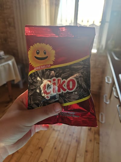 A hand holds a red, foil-wrapped bag of "Çiko" brand sunflower seeds, with the word "PREMIUM" above a cartoon sunflower with a smiling face. The bag displays an image of numerous black sunflower seeds. The background suggests an indoor setting with wooden flooring, a table covered with a tablecloth, and a bright window with sheer curtains. The scene appears to be a casual moment, possibly in a home environment. The image does not provide any specific information about the city of Saray, Azerbaijan.
