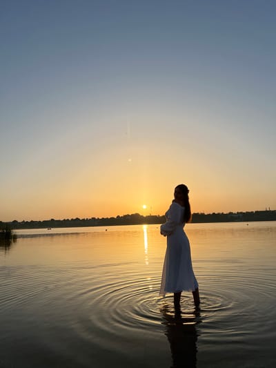 The image captures a silhouette of a woman standing in shallow water at sunset. The woman is wearing a white dress and is facing away from the viewer, looking towards the setting sun. The water is calm, with subtle ripples reflecting the warm hues of the sky. The background features a tree-lined horizon and a faint outline of buildings, suggesting an urban or semi-urban environment. The sky is a gradient of soft blues and oranges, characteristic of a sunset.
This scene is likely located at the Ternopil Lake in Ternopil, Ukraine. Ternopil Lake is a well-known reservoir located within the city, surrounded by parks and urban areas. The presence of a calm body of water, a tree-lined shore, and hints of buildings in the background are consistent with the landscape of the Ternopil Lake embankment. The image depicts a moment of serene contemplation at dusk, with the natural beauty of the sunset enhanced by the silhouette of the solitary figure.