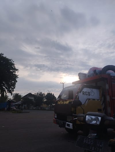 This image captures an outdoor scene at what appears to be either sunrise or sunset, indicated by the low, diffused light filtering through a cloudy sky. The setting is Kecamatan Jangkar, Indonesia.
The primary subjects are a yellow and red dump truck and the front portion of a black motorcycle, both parked. The truck, facing slightly left, has "RGS" written on its side window, accompanied by a hand gesture that resembles a peace sign or "V" sign. Its bed is loaded with items covered by tarps and possibly some spare tires. The motorcycle, positioned in the lower right foreground, obscures part of the truck's front bumper. Its license plate, clearly visible, reads "P 6855 WJ" and "12-19," consistent with an Indonesian vehicle registration.
In the mid-ground, to the left of the truck, there's an open, asphalted area, possibly a parking lot or a wide road. Further back, utility poles with power lines stretch across the frame, and glimpses of residential buildings with dark roofs are visible amongst scattered trees. The foliage on the left side of the frame, belonging to a large tree, is dark against the brighter sky.
The sky is largely overcast with stratocumulus or altocumulus clouds, through which the sun emits a soft, warm glow. A very small, dark, elongated object (possibly a bird or insect) is visible high in the sky in the upper left quadrant. No specific activity or event is taking place; the scene appears tranquil and still. The overall mood is calm, with the cloudy conditions and low light contributing to a subdued atmosphere.