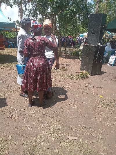Three women are standing in a grassy area in Malava, Kenya (latitude: 0.519, longitude: 34.81). Two of the women are wearing brightly colored head wraps and the third woman is wearing a hat. They are all facing the camera. The woman in the center is wearing a long, brown dress with a floral pattern. She has her hands clasped in front of her. The woman on the left is wearing a white long-sleeved shirt with a white hat. She has a blue bag strapped to her wrist. The woman on the right is wearing a white shirt with a white head wrap. She has her hand on the woman in the center's shoulder. There are several people in the background, but they are out of focus. A large, black speaker is located in the background to the right of the women. The scene is in a rural area. The ground is dirt and there is a lot of dry grass on the ground. The sky is clear.