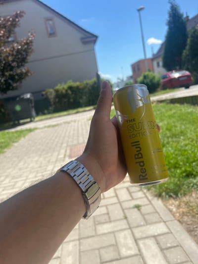 The image shows a hand holding a yellow can of Red Bull "Summer Edition" energy drink. The can has text on it, including "THE SUMMER EDITION" and "Red Bull". The background is blurred but shows a paved walkway, green grass, and parts of buildings, suggesting an outdoor setting during the daytime. The sun is shining, indicated by the bright lighting and clear blue sky visible in the upper left portion of the image. The overall atmosphere appears casual and summery.