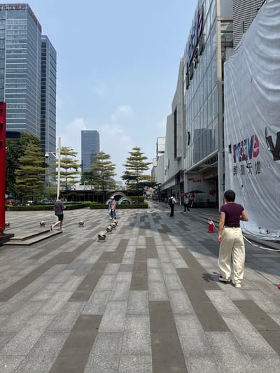 A street scene in Tianhe District, China, shows a wide, paved pedestrian walkway lined with modern buildings and trees under a clear sky. In the foreground, a woman with short dark hair wearing a purple t-shirt and loose cream-colored pants stands with her back to the camera, facing away from the viewer and towards the buildings. To her left, a man in a dark t-shirt and shorts walks away from the camera. Further down the walkway, another person in a bright orange and blue vest is walking. The buildings on the right are a mix of glass-fronted structures, with one large building featuring a white banner with colorful lettering. On the left, tall, dark skyscrapers with reflective windows dominate the background, interspersed with lush green trees. Several small, round, yellow and black bollards are visible along the walkway. Traffic is visible in the distance on the left.