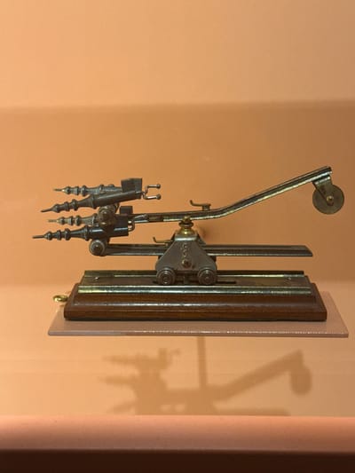 This media file is a photograph of an antique mechanical instrument mounted on a wall. The instrument appears to be made of metal and wood, with several articulated arms and a roller at the top. It is positioned against a peach-colored wall, and a shadow of the instrument is cast on the wall behind it. The instrument has a complex, almost skeletal appearance with various levers, pivots, and what seem to be drawing points or styluses at the bottom.
The setting appears to be indoors, likely a museum or a historical display given the nature of the object and its presentation. There are no people visible in the photograph. The lighting suggests it's daytime, but the artificial lighting setup is not discernible. The object itself is the sole subject of the photograph.
Notable details include the intricate craftsmanship of the mechanical device, suggesting it might be an early drafting tool, a musical automaton component, or a scientific instrument. The wooden backing board is rectangular with rounded corners, and a small brass knob is visible at the bottom right of the board. The metallic parts of the instrument show signs of age and use, with a patina that hints at its historical significance. The shadow on the wall is detailed, mirroring the shape of the instrument and further emphasizing its three-dimensional form. No visible text is present on the instrument or the wall. The overall impression is one of historical preservation and the appreciation of intricate machinery.