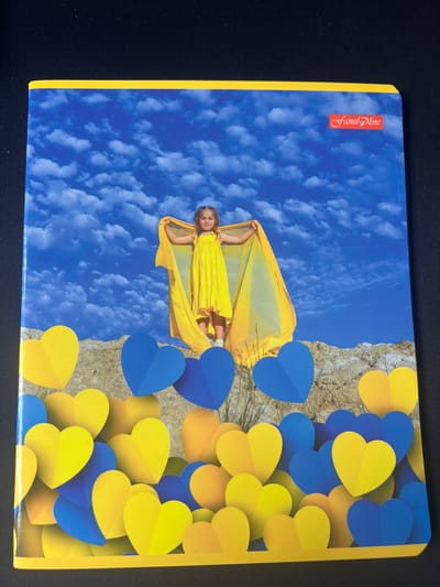 This media file is a cover of a notebook. The cover features a young girl in a yellow dress and a yellow cape, standing on a rocky cliff against a bright blue sky with fluffy white clouds. She is holding her cape outstretched as if she is flying. Numerous blue and yellow paper hearts are arranged in a pattern, partially obscuring the cliff and the sky. The hearts create a vibrant, layered effect. The yellow edge of the notebook is visible. A red logo with white text that reads "FamilDine" is located on the top left of the cover. The overall impression is cheerful and uplifting, with a strong visual theme of yellow and blue. The scene suggests a bright, sunny day.