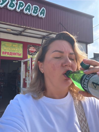 A woman with blonde hair is drinking from a green glass bottle of mineral water in front of a store. The store has a red facade and a sign that says "УБРАВА" in large white letters with a green outline. Below this sign, there is a smaller sign in Russian that says "ООО "МИГ" МАГАЗИН №11 ПРОДУКТЫ", indicating it is a store named "MIG" and sells groceries. The bottle has a label that reads "ИСТОЧНИКИ КАВКАЗА" and a number "4". The woman is wearing a white sweatshirt and has a strap with a geometric pattern across her chest. The scene appears to be outdoors on a sunny day. Leningradskaya, Russia is a town in Krasnodar Krai, but there is no specific visual indicator to confirm the location beyond the text on the signs.