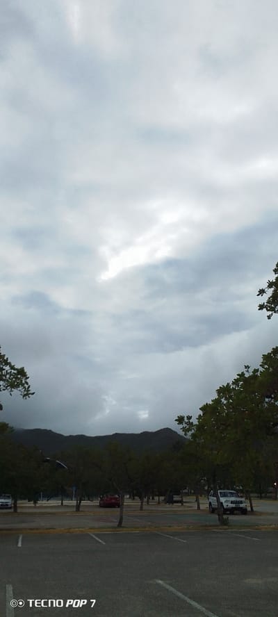 The image depicts an outdoor scene with a paved parking lot in the foreground. Several trees with green foliage are visible, framing the upper corners of the image. In the background, a dark, mountainous landscape is shrouded in a cloudy sky, with a break in the clouds allowing some light to filter through. A few cars are parked in the lot, including a white SUV and a red car. The scene appears to be in a natural setting, possibly on the outskirts of a city. The text "TECNO POP 7" is watermarked on the bottom left of the image. Without further context, it is not possible to definitively pinpoint the exact location within Pedro González, Venezuela.
